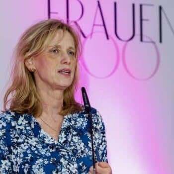 Frau mit blonden Haaren spricht am Mikrofon vor einem pinken Hintergrund mit dem Text "FRAUEN 100".