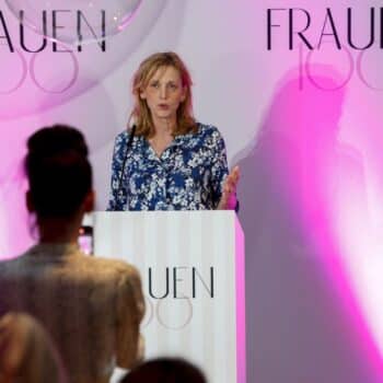 Frau spricht am Rednerpult vor einem Banner mit dem Schriftzug "FRAUEN 100", während eine andere Person sie fotografiert.