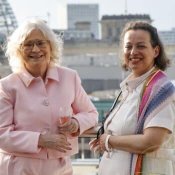 Zwei Frauen stehen lächelnd auf einer Terrasse mit Stadtansicht, eine hält ein Glas Sekt.