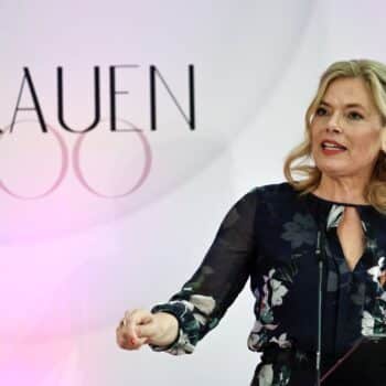 Frau mit blonder Haarfrisur spricht am Mikrofon vor einer Wand mit dem Schriftzug "FRAUEN 100".