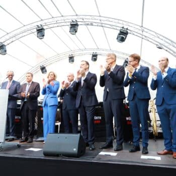 Gruppierung von Menschen auf einer Bühne, die applaudieren, mit Mikrofon und Musikinstrumenten im Hintergrund.