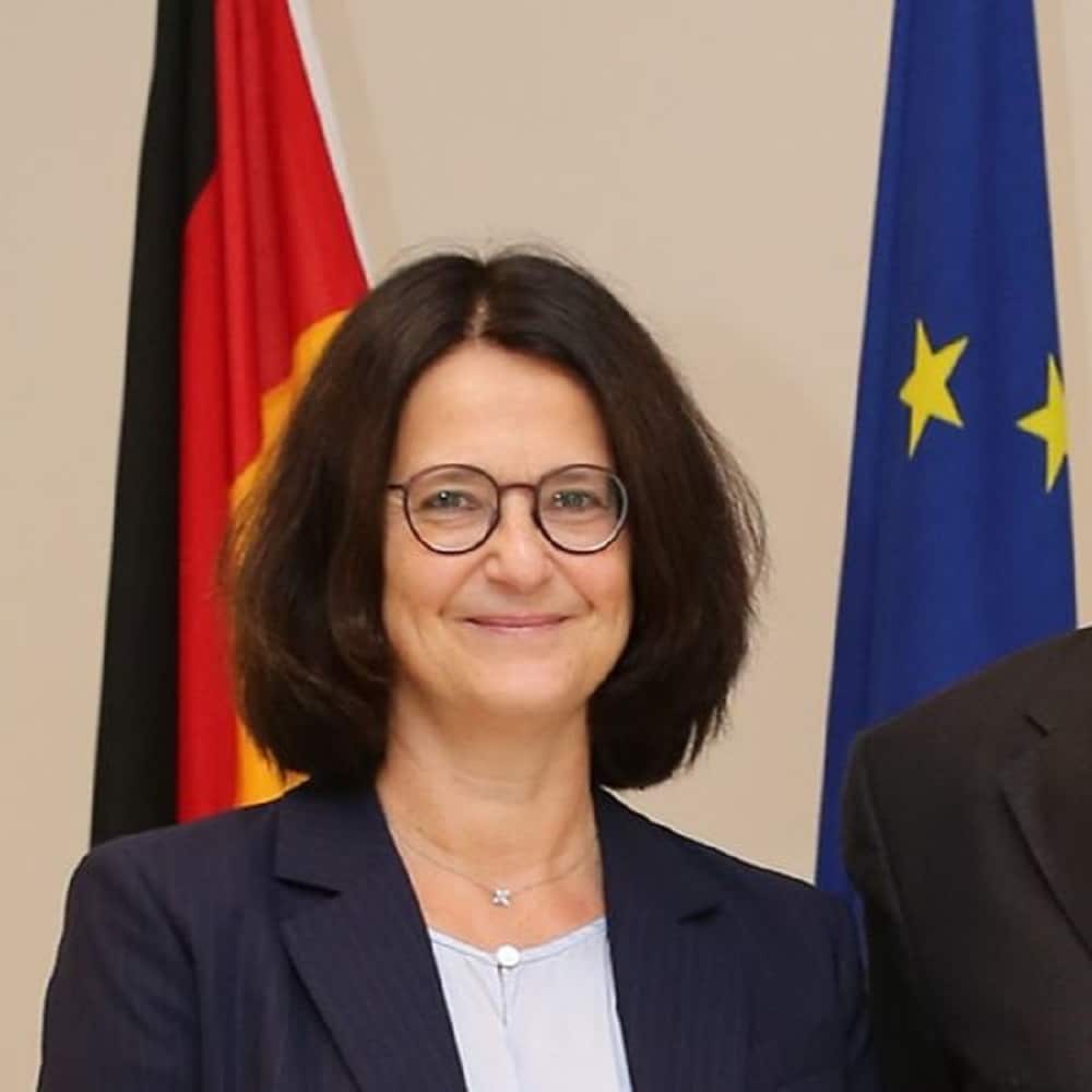 Porträt einer Frau mit Brille, lächelnd, vor Flaggen Deutschlands und der EU.