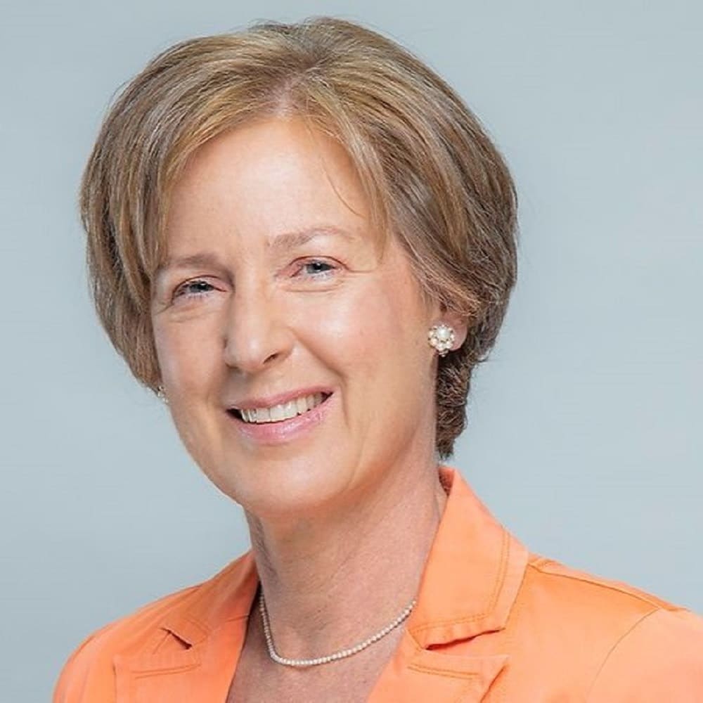 Porträt einer lächelnden Frau mit kurzen, blonden Haaren, die ein orangefarbenes Jackett trägt. Heller Hintergrund.