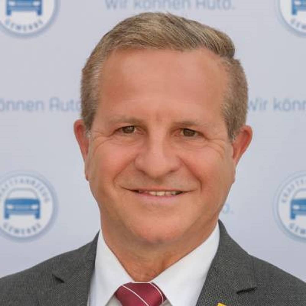 Porträt eines Mannes mit kurzen, grauen Haaren, Anzug und Krawatte, lächelnd vor einem hellen Hintergrund mit Automotiven.