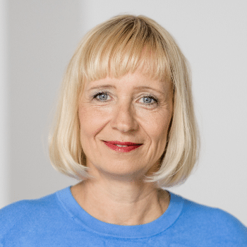 Porträt einer blonden Frau mit schulterlangen Haaren, blauer Bluse und freundlichem Lächeln vor neutralem Hintergrund.