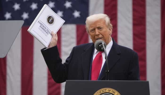 Donald Trump spricht am Mikrofon und hält ein Dokument in der Hand, vor einer US-Flagge im Hintergrund.