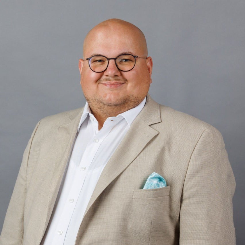 Mann mit Glatze und Brille, trägt einen hellen Anzug und ein weißes Hemd, lächelt vor grauem Hintergrund.