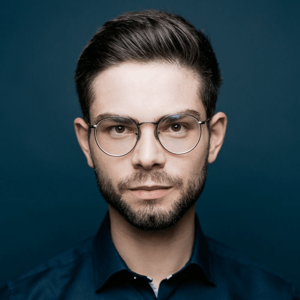 Porträt eines jungen Mannes mit Brille, dunklem Hemd und neutralem Gesichtsausdruck vor einem blauen Hintergrund.
