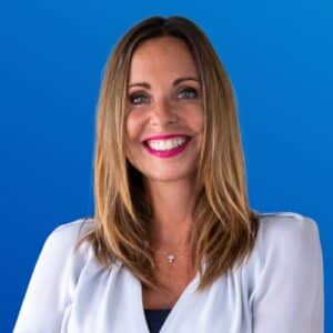 Frau mit langen, blonden Haaren und rotem Lippenstift lächelt vor blauem Hintergrund.