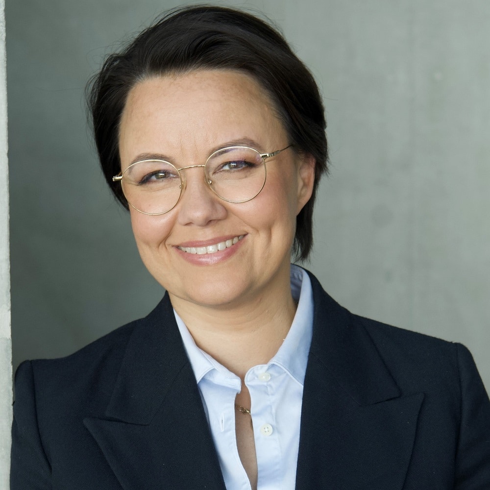 Porträt von Michelle Müntefering mit Brille, lächelnd, in einem blauen Blazer vor einer grauen Wand.