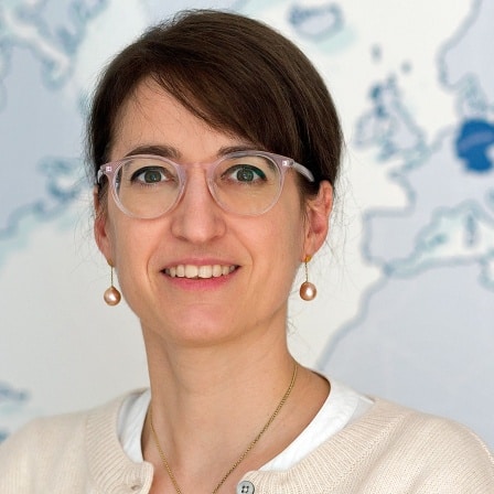 Porträt einer Frau mit Brille, lächelnd vor einer Weltkarte im Hintergrund. Sie trägt eine helle Bluse und eine Kette.