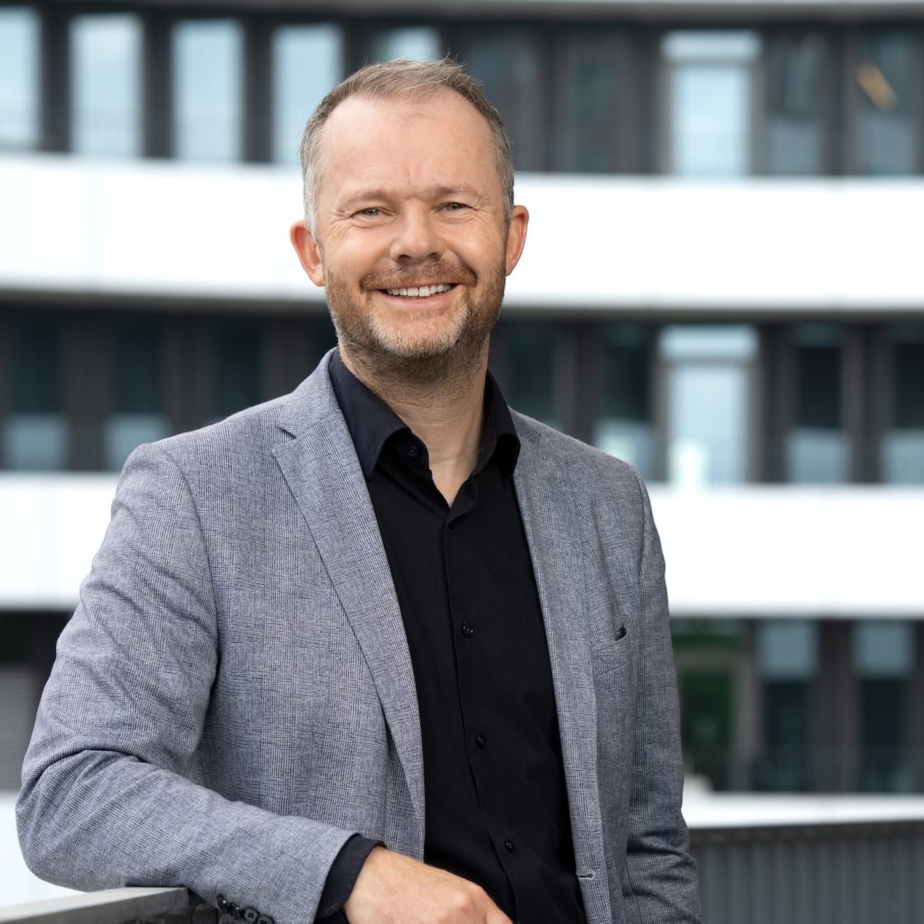 Mann in grauem Blazer lächelt vor modernem Gebäude mit Fenstern.