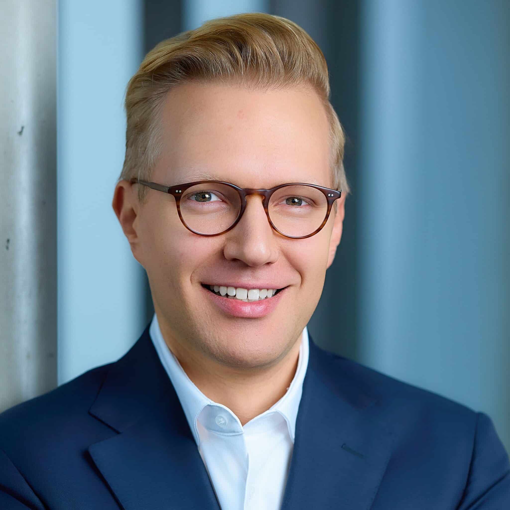 Porträt eines lächelnden Mannes mit Brille, blonden Haaren, in einem dunkelblauen Anzug vor einem hellen Hintergrund.