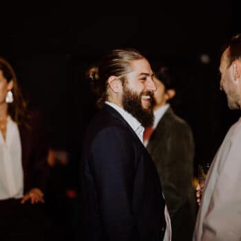 Zwei Männer im Gespräch, umgeben von weiteren Personen bei einem Event, mit Getränken in der Hand.