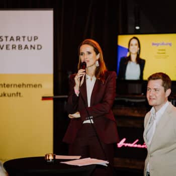 Frau mit Mikrofon spricht auf einer Veranstaltung, neben ihr steht ein Mann. Im Hintergrund ein Bildschirm mit Präsentation.