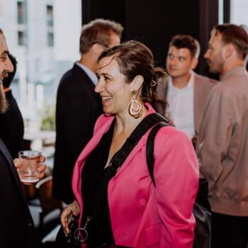 Menschen in formeller Kleidung unterhalten sich auf einem Event, eine Frau trägt einen pinken Blazer und lächelt.