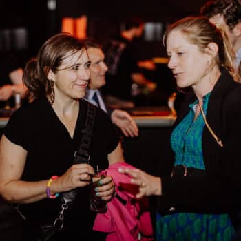 Zwei Frauen unterhalten sich lächelnd bei einem Event, im Hintergrund sind weitere Personen und eine Bar zu sehen.