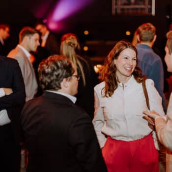 Menschen in formeller Kleidung unterhalten sich bei einem Event, mit bunten Lichtern im Hintergrund.