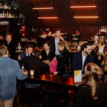 Menschen in einem stilvollen Bar-Setting, die sich unterhalten und Getränke genießen, mit dimmiger Beleuchtung und Pflan...