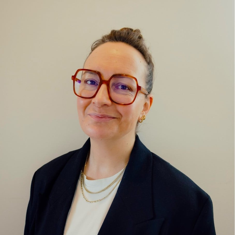 Frau mit Brille und Hochsteckfrisur, trägt einen schwarzen Blazer und eine Kette, lächelt vor neutralem Hintergrund.