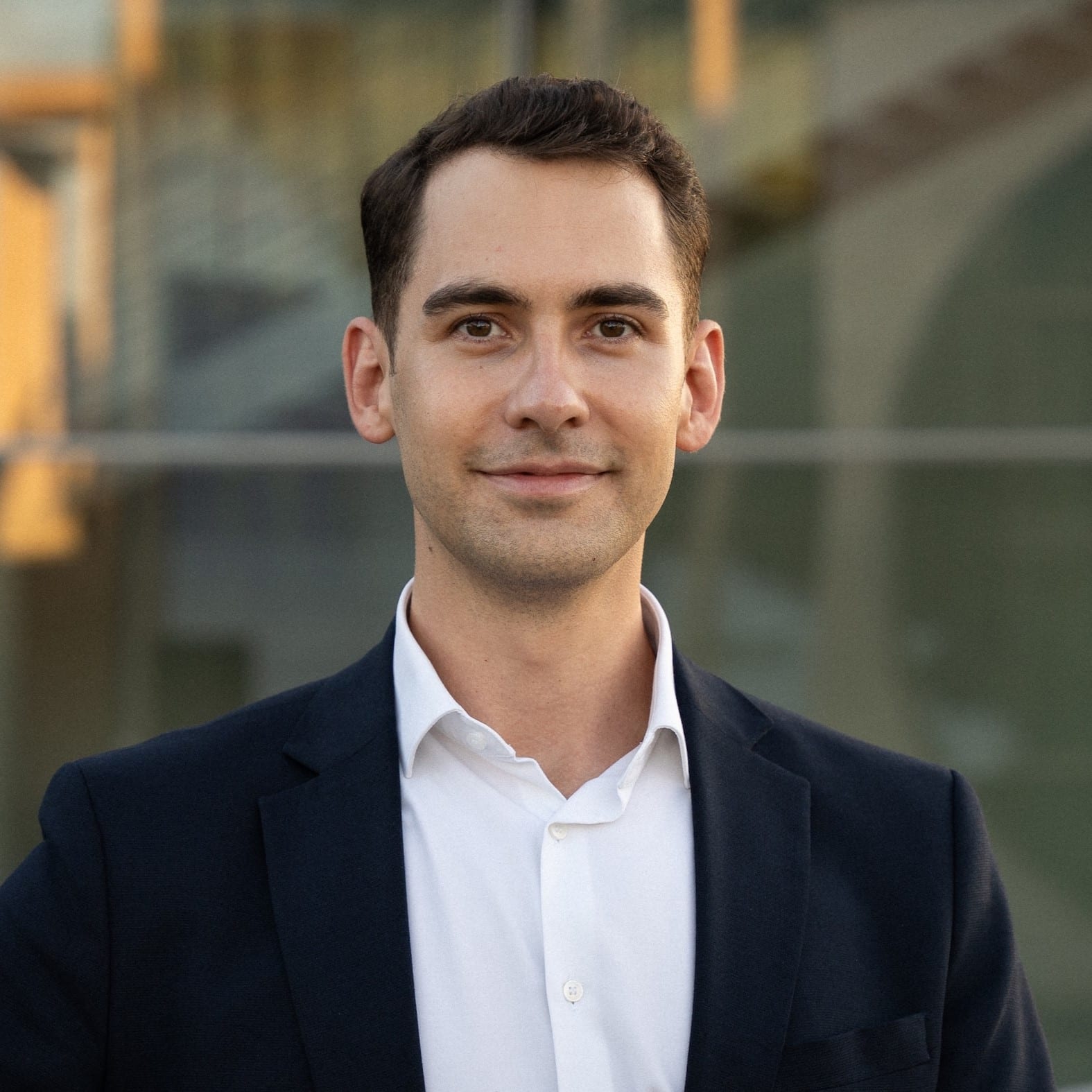 Porträt eines Mannes in einem Anzug, der lächelt und vor einem modernen Gebäude steht.