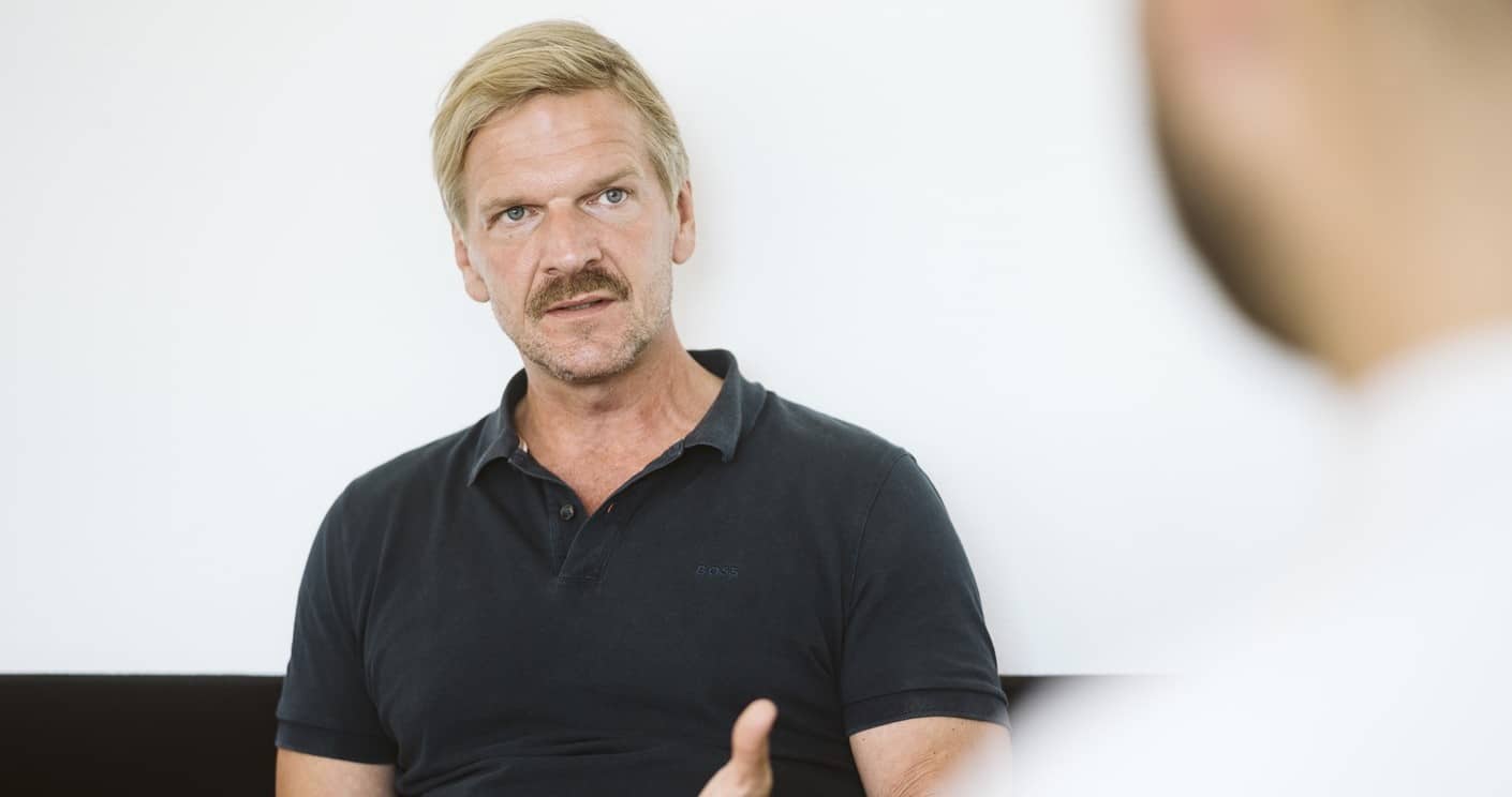 Mann mit blonden Haaren und Schnurrbart spricht ernsthaft, im Hintergrund ist eine unscharfe Person zu sehen.