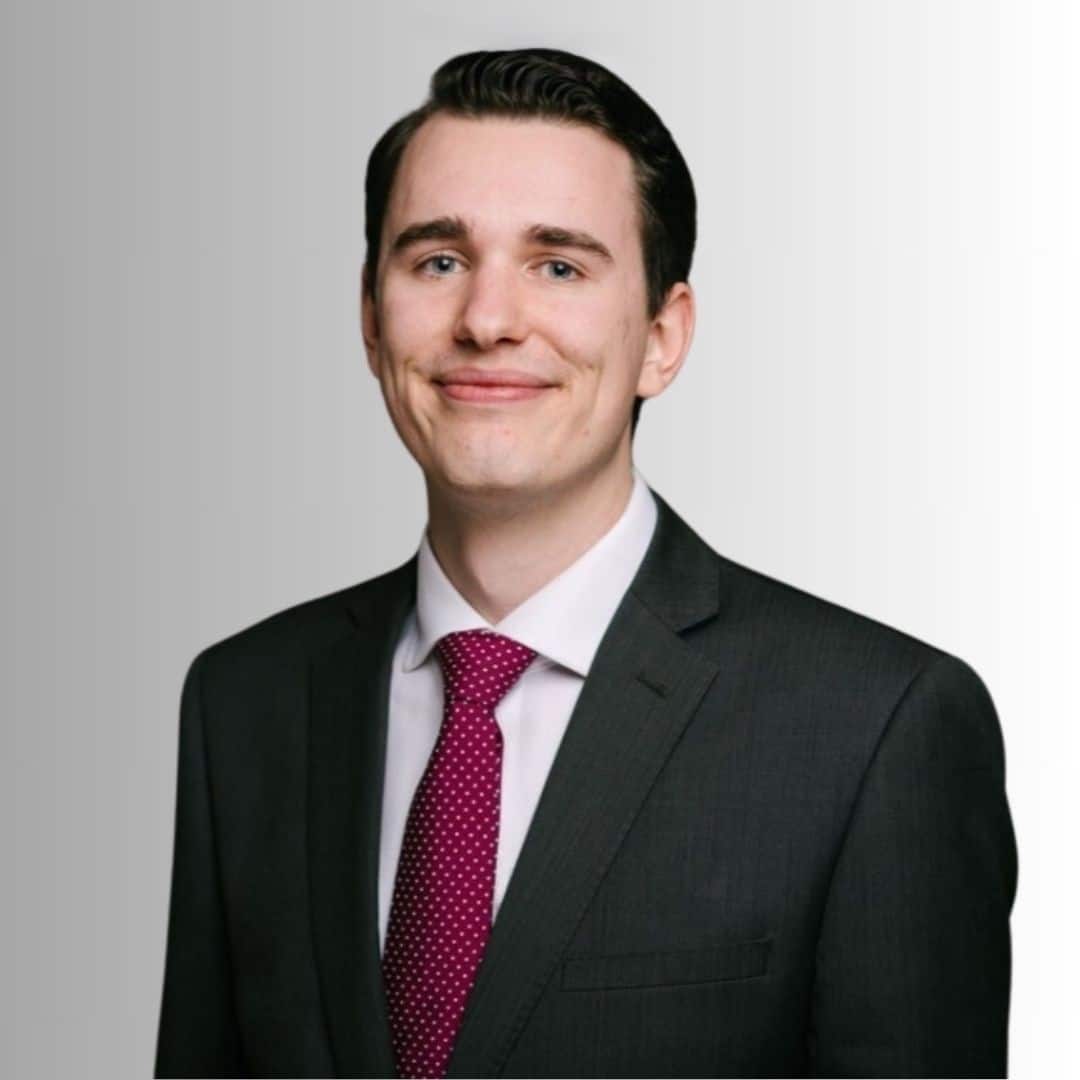 Junger Mann in Anzug mit Krawatte, lächelt vor einem hellen, einfarbigen Hintergrund.