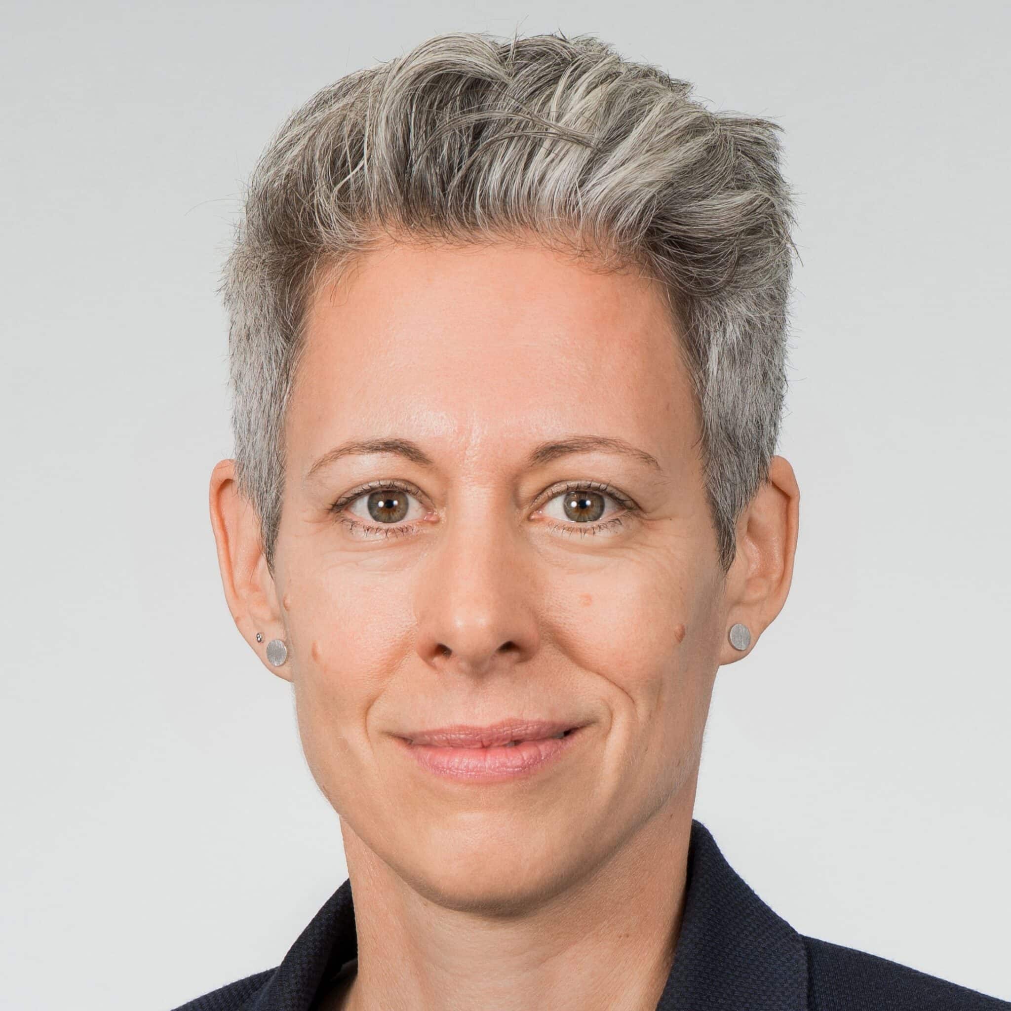 Porträt einer Frau mit kurzen grauen Haaren, neutralem Gesichtsausdruck, in einem dunklen Blazer vor hellem Hintergrund.