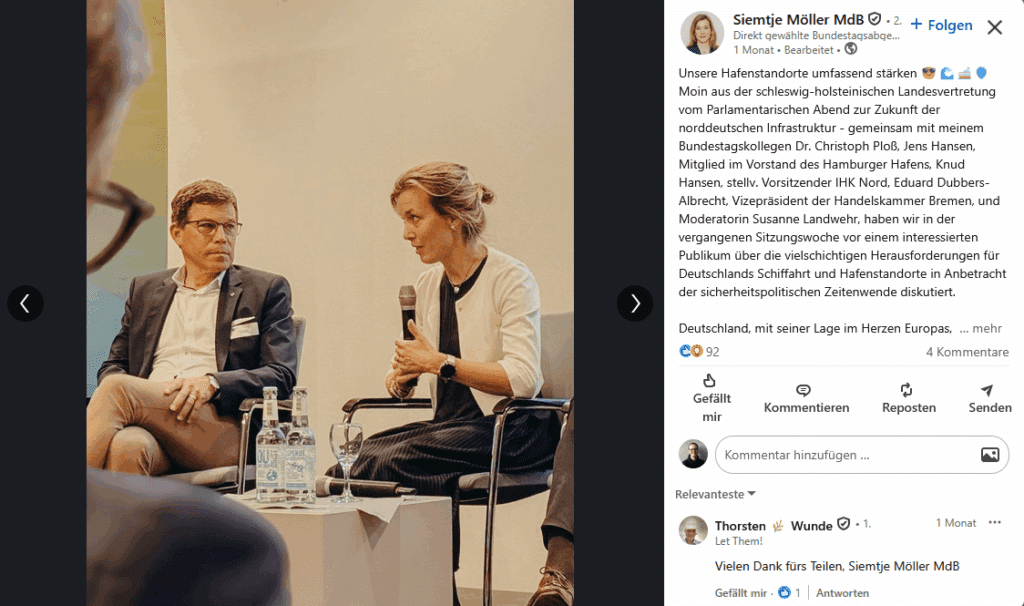 Zwei Personen sitzen auf einer Bühne, während eine von ihnen in ein Mikrofon spricht. Im Hintergrund sind Publikum und G...