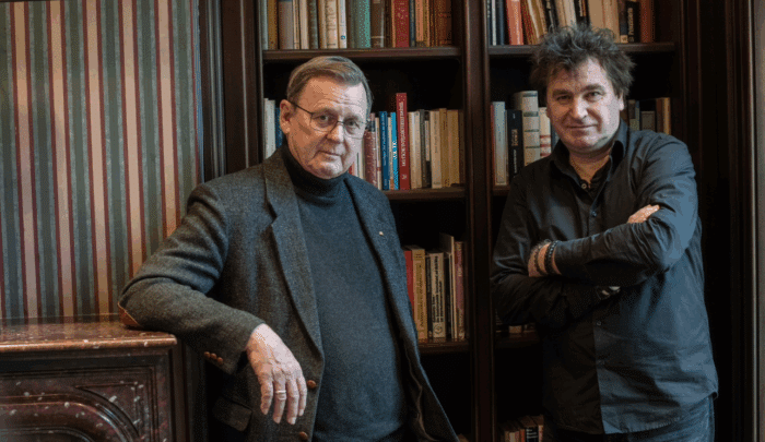 Zwei Männer stehen in einem Raum mit Bücherregalen, einer in einem grauen Anzug, der andere in schwarzer Kleidung.