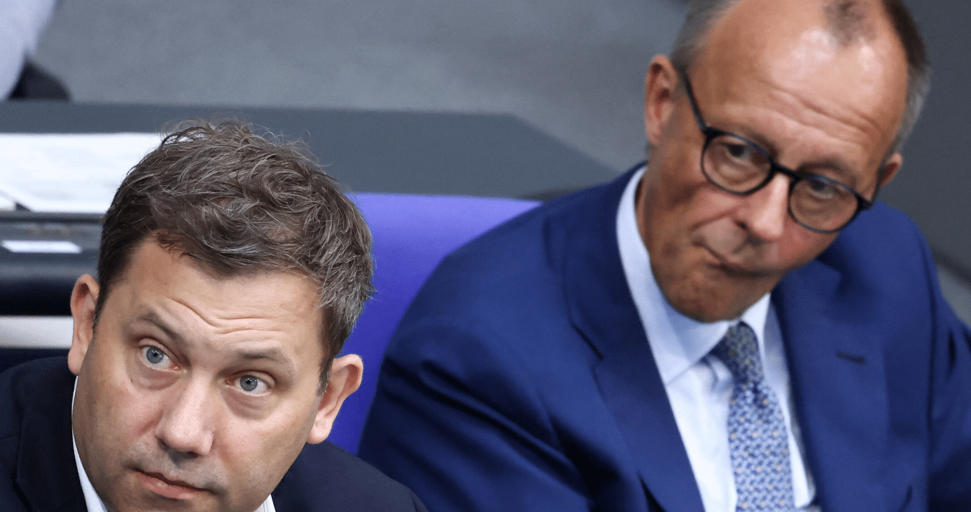 Zwei Männer in Anzügen sitzen im Plenarsaal, einer schaut nachdenklich, der andere wirkt skeptisch.