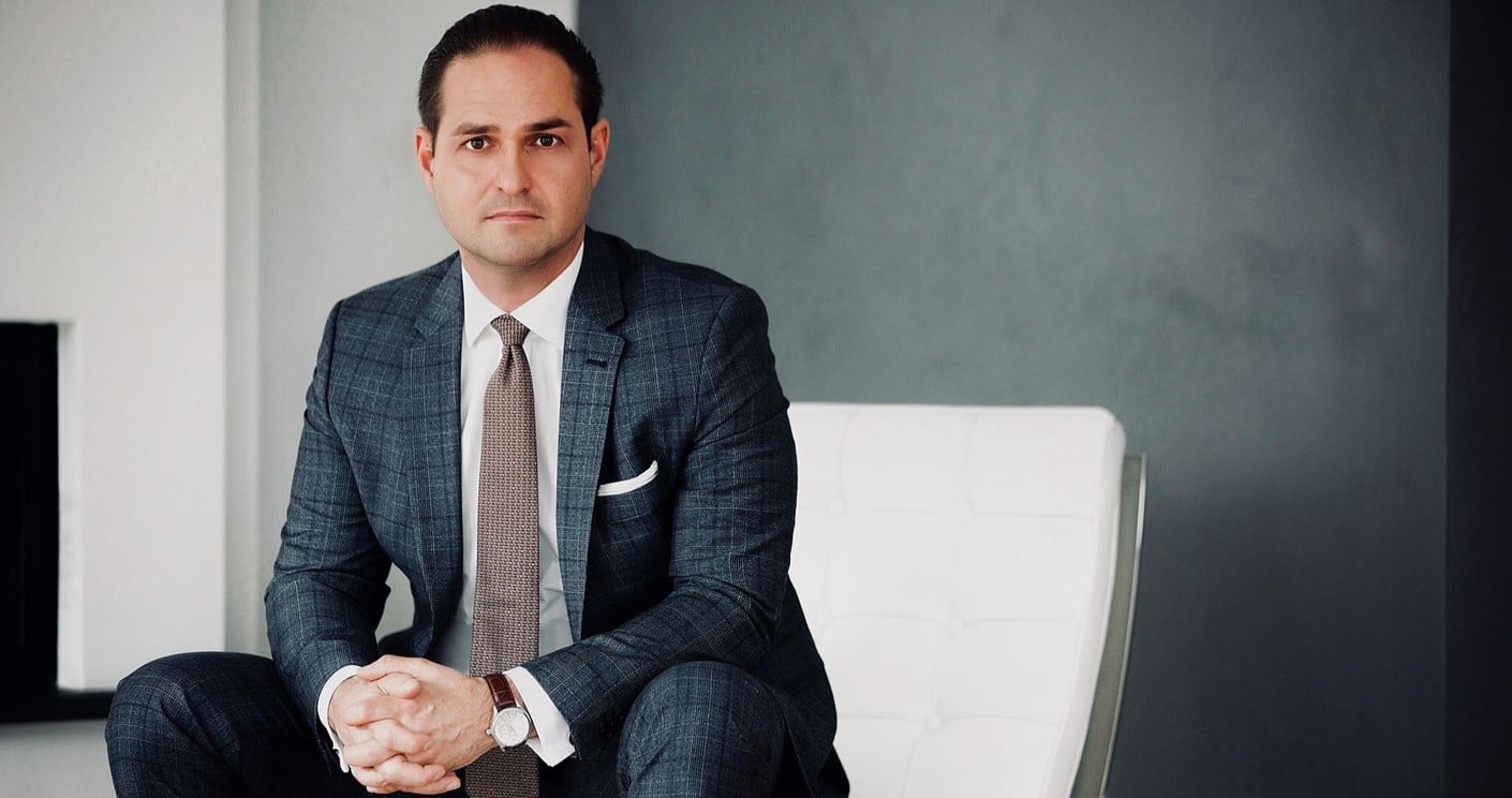 Mann in Anzug sitzt auf einem weißen Stuhl vor grauem Hintergrund, mit ernstem Blick und gefalteten Händen.