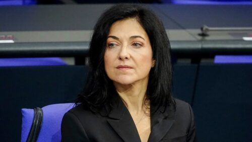 Frau mit schwarzem Haar und Anzug sitzt nachdenklich in einem Sitzungssaal mit blauen Stühlen im Hintergrund.