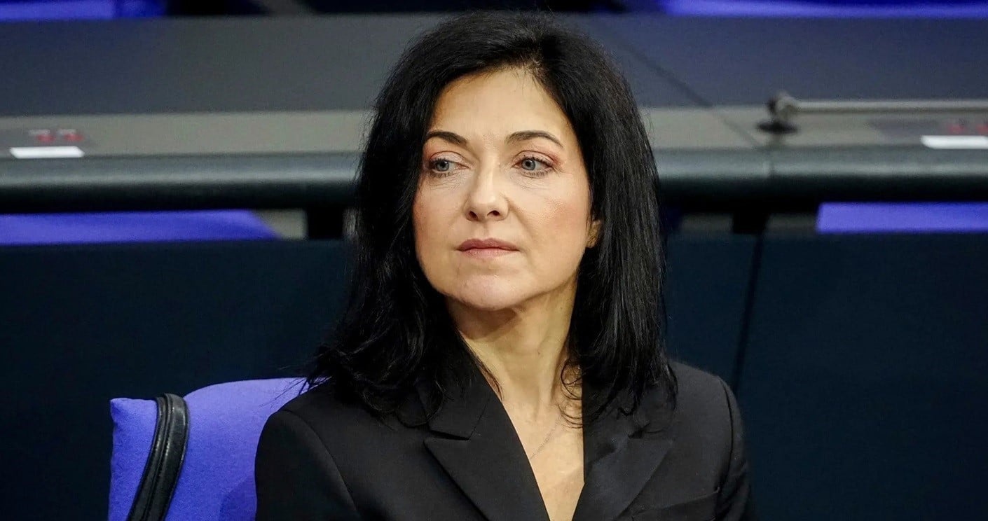 Frau mit schwarzem Haar und Anzug sitzt nachdenklich in einem Sitzungssaal mit blauen Stühlen im Hintergrund.