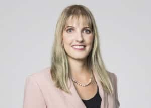 Porträt einer lächelnden Frau mit blonden Haaren, in einem blauen Oberteil und einem rosa Blazer, vor neutralem Hintergr...