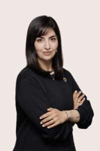 Porträt einer Frau mit langen, dunklen Haaren, die in einem schwarzen Pullover mit verschränkten Armen lächelt.