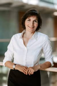 Frau mit kurzer brauner Haarfrisur, trägt ein weißes Hemd und steht an einem Geländer in einem modernen Innenraum.