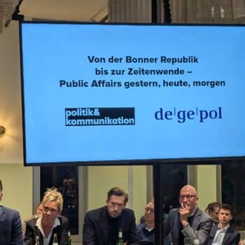 Podiumsdiskussion mit mehreren Personen, einem Bildschirm im Hintergrund mit dem Titel der Veranstaltung und Logos.