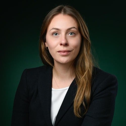 Porträt einer Frau mit langen, braunen Haaren, in schwarzer Blazerjacke, vor einem dunklen, grünen Hintergrund.