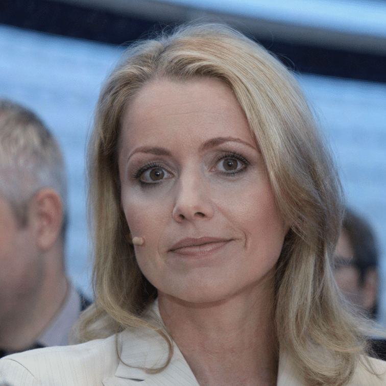 Frau mit blonden Haaren und Mikrofon, schaut direkt in die Kamera, im Hintergrund verschwommene Personen.