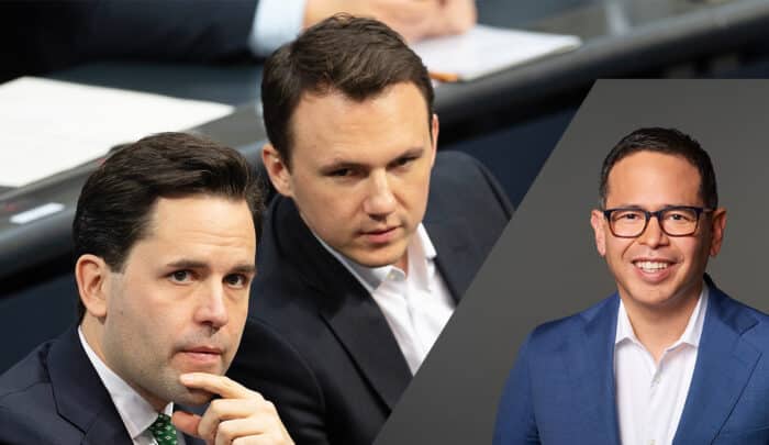 Zwei Männer in Anzügen sitzen an einem Tisch, während ein dritter Mann in einem blauen Anzug lächelt.