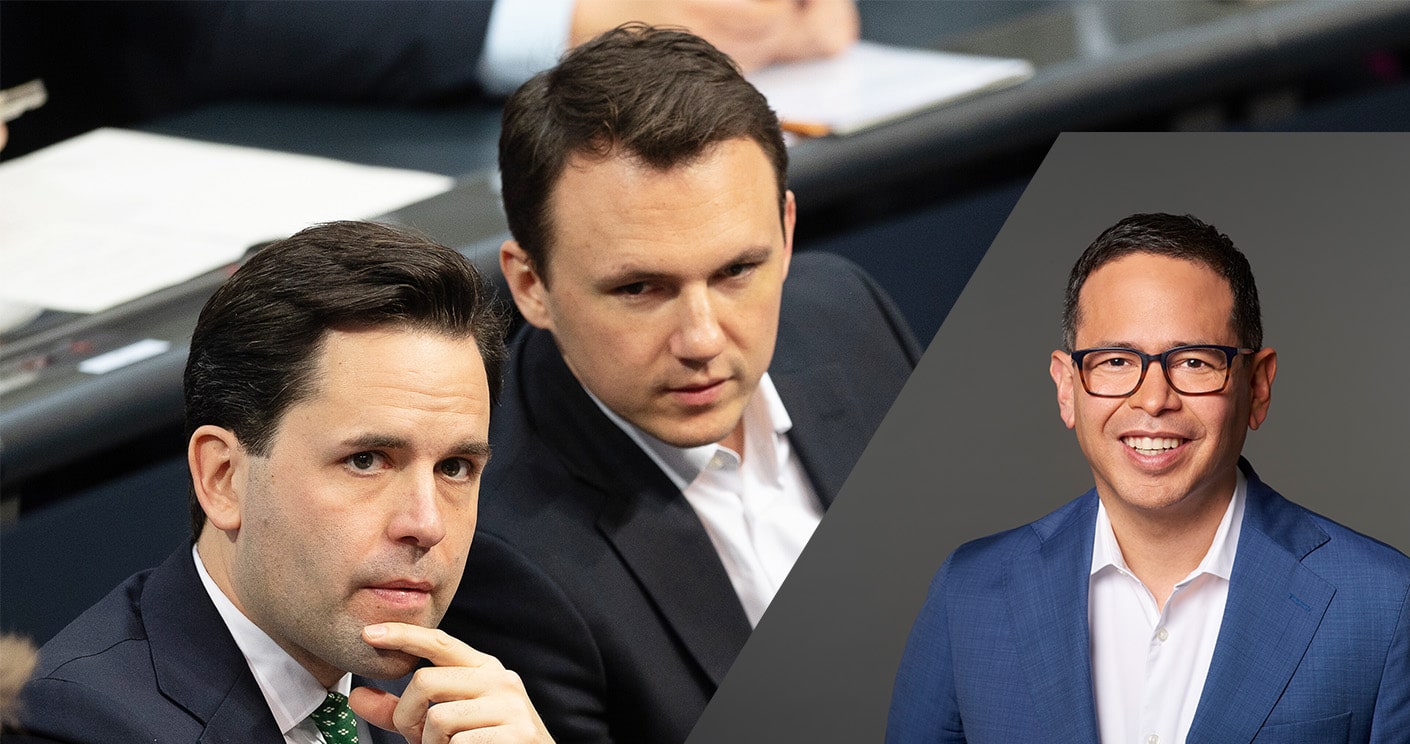 Zwei Männer in Anzügen sitzen an einem Tisch, während ein dritter Mann in einem blauen Anzug lächelt.