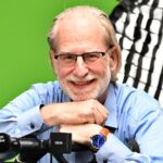 Mann mit Brille und grauem Bart lächelt, stützt sich auf einen Mikrofonständer, grüne Hintergrundfarbe.