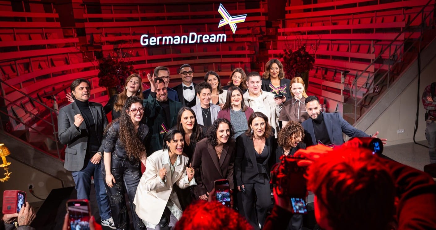 Gruppenfoto von Menschen bei einer Veranstaltung mit dem Schriftzug "German Dream" im Hintergrund. Rotes Licht beleuchtet ...