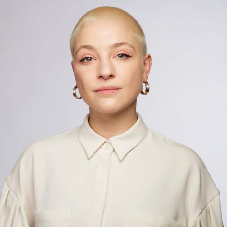 Porträt einer Person mit kurzem, blonden Haar, die ein helles Hemd trägt und in die Kamera schaut.