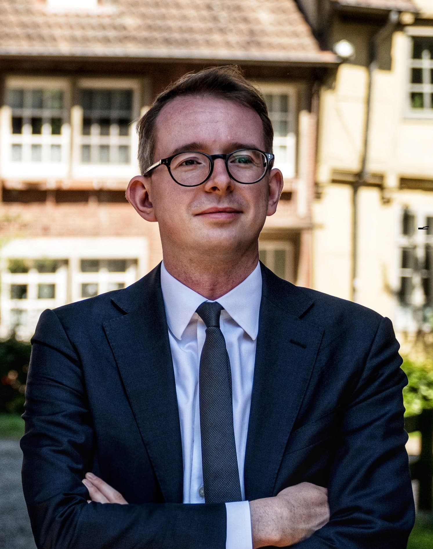 Mann in Anzug mit Brille steht mit verschränkten Armen vor einem Gebäude mit Fenstern.