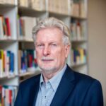 Peter Voß. Foto: Moritz Vennemann Mann mit grauem Haar in Anzug steht vor einem Bücherregal mit bunten Buchrücken.