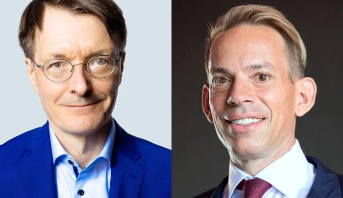 Zwei Männer im Anzug, einer mit Brille und blauer Jacke, der andere mit hellen Haaren und roter Krawatte, nebeneinander.