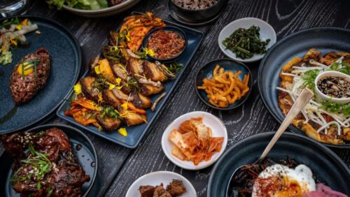 Foto: Unsplash / Alexandra Tran Vielfalt an asiatischen Gerichten auf dunklen Tellern, darunter Fleisch, Gemüse, Reis und Beilagen.