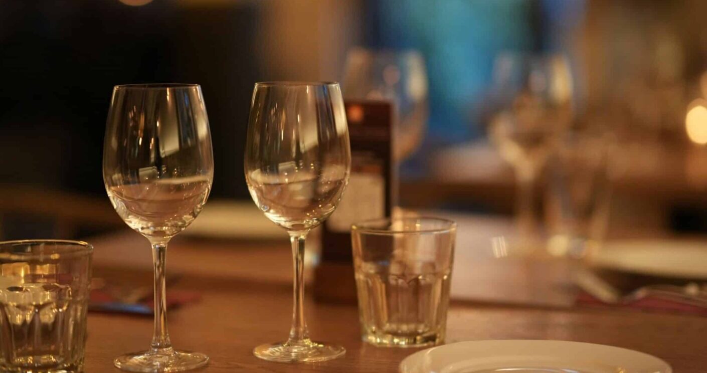 Zwei Weingläser und ein Wasserglas auf einem gedeckten Tisch, sanfte Beleuchtung im Hintergrund.